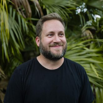 Author photo for P. Scott Cunningham. Author wears black T-shirt and is standing in front of a palm frond.